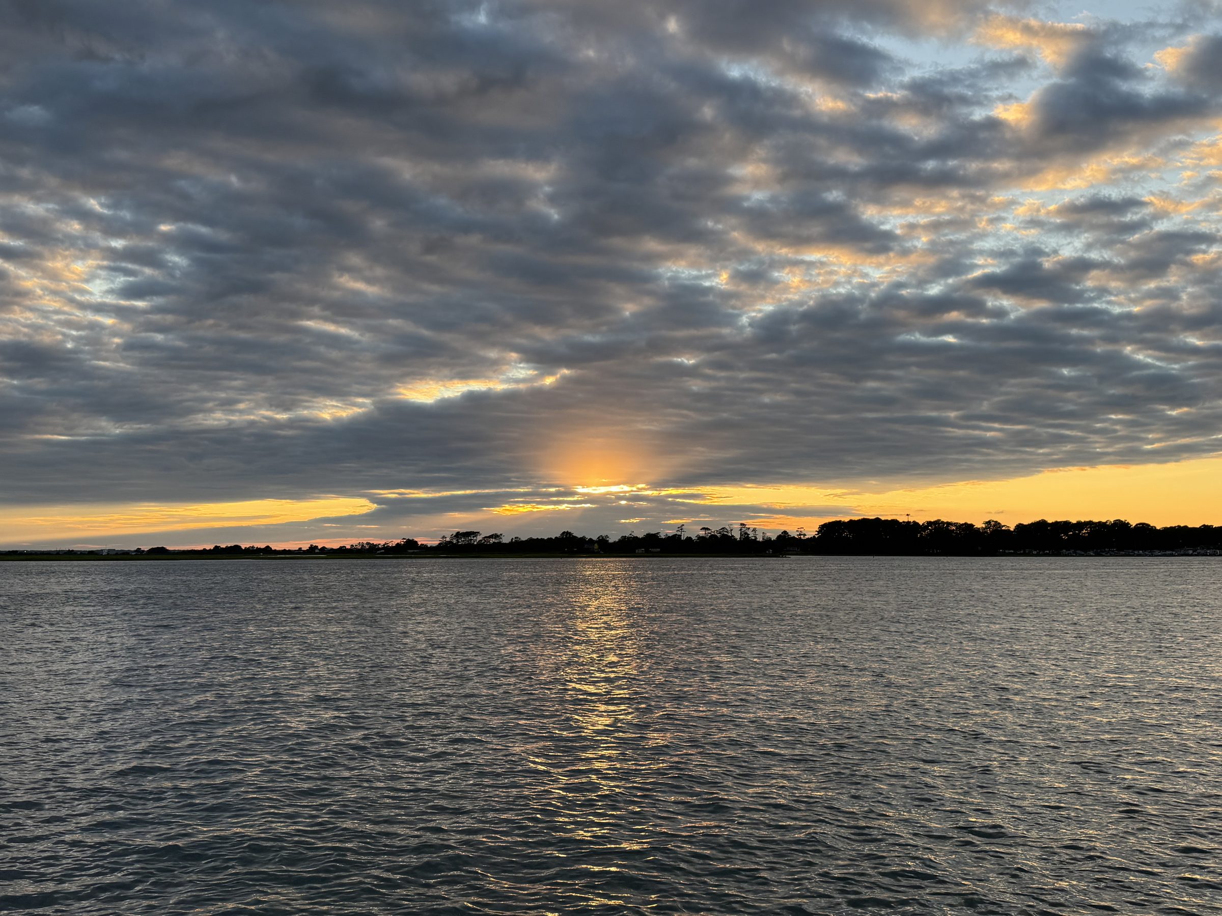 Sunset over the water at Assateague Island, the sun breaking through banded clouds above the tree line.